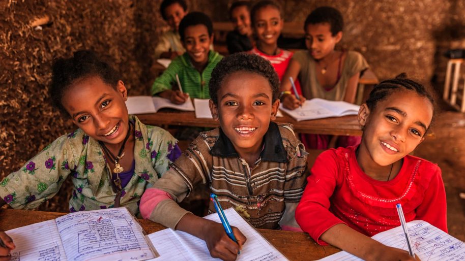Des enfants sourient à leur pupitre, cahiers ouverts, dans une salle de classe.